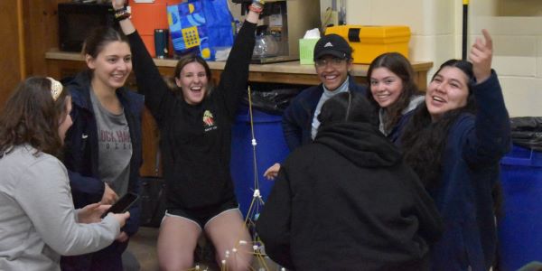 Jenna with her fists pumped in front of a spaghetti tower, surrounded by her teammates and friends